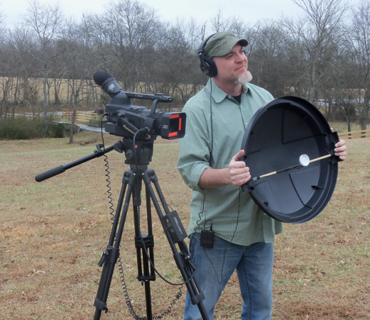 Shot of videographer holding a DIY parabolic mic dish, standing next to a camcorder on a tripod.